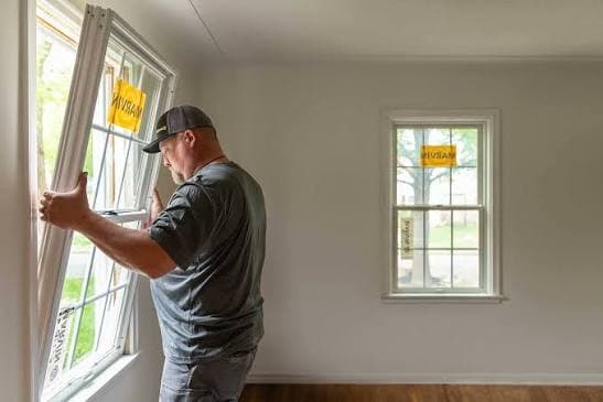 A Black Bear installer fitting a new double-hung window in a freshly painted room, with another new window across the room